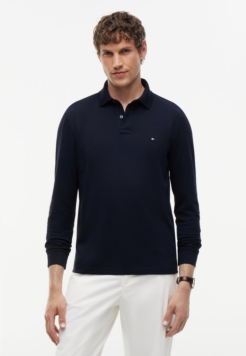 Hombre con una camiseta polo azul marino de manga larga y pantalones blancos, de pie contra un fondo claro y liso.