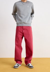 Szara bluza z okrągłym dekoltem i białym logo na środku, zestawiona z luźnymi, czerwonymi dżinsami i czarno-białymi trampkami.