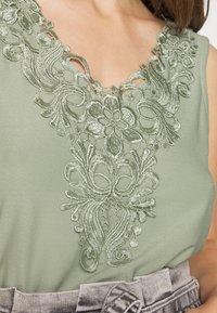 Gros plan sur un haut sans manches couleur vert sauge avec une broderie en dentelle florale complexe au niveau de l'encolure, assorti à un pantalon taille haute gris délavé.