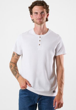 T-shirt blanc à manches courtes côtelé avec trois boutons marron au col, associé à un jean en denim bleu. Présente une coupe décontractée.