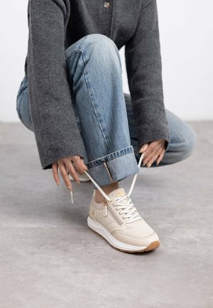 Zapatilla de color beige claro con una parte superior texturizada, acentos dorados y cordones blancos. La suela está acolchada con una base marrón para mayor tracción.
