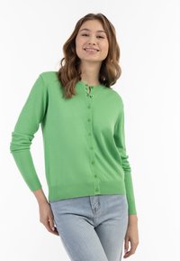 Žalias kardiganas su minkšta tekstūra, apvaliu iškirpimu ir mygtukais per visą priekį. Turi ilgus rankovėmis ir nėriniuotą apykaklę bei rankoves.