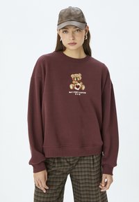 Sudadera marrón con un gráfico de un oso y texto que dice "PERO PRIMERO CAFÉ". Presenta puños y dobladillo acanalados. Combinada con pantalones de cuadros.