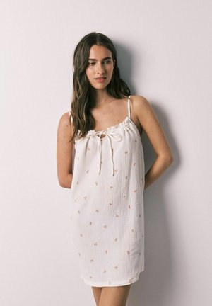 Vrouw met lang bruin haar die een witte mouwloze jurk met kleine bloemenprints draagt, staat tegen een lichte muur met haar handen achter haar rug.