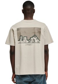 Beżowa bawełniana koszulka z grafiką przedstawiającą budynki i góry na plecach, z czarnymi i brązowymi akcentami oraz nakładkami tekstowymi.