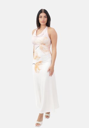 Mujer con cabello largo y oscuro, vestida con un vestido blanco sin mangas con estampados florales en melocotón y sandalias blancas de tacón, de pie sobre un fondo blanco.