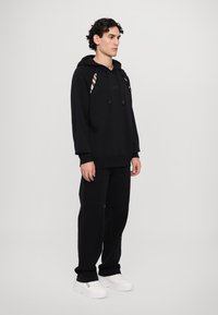 Sweatshirt noir à capuche avec cordons, poche kangourou et logo ; associé à un pantalon noir et des baskets blanches épaisses. Design simple.