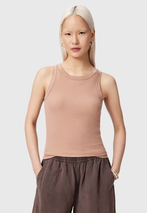 Jeune femme aux cheveux plats blond platine, vêtue d'un haut beige sans manches et d'un pantalon marron ample, debout les mains dans les poches.