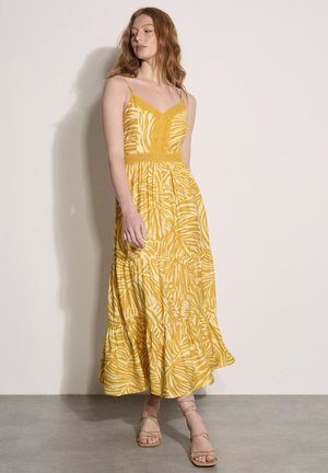 Kobieta ubrana w żółtą, wzorzystą, bezrękawnikową sukienkę maxi z koronkowymi detalami, stojąca przy gładkiej beżowej ścianie i szarej podłodze.