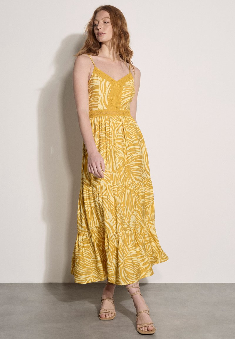 Femme portant une robe longue sans manches jaune à motifs avec des détails en dentelle, debout devant un mur beige uni et un sol gris.