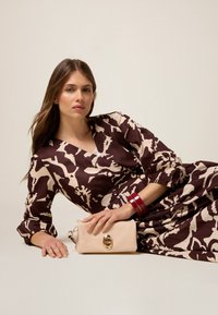 Abito a motivi marroni e beige con maniche voluminose, abbinato a una clutch beige chiaro con un accento in hardware dorato.