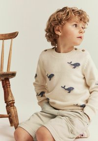 Strik en cremede sweater med hvalmønstre i marineblå, træknapper på skulderen. Kombination med beige shorts, siddende på en træstol.