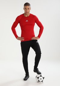 Rode lange mouwen sportshirt met een strakke pasvorm en Adidas-logo; gecombineerd met zwarte tapered broek met gouden strepen en een voetbal.