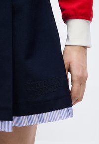 Χέρι ακουμπισμένο δίπλα σε σκούρα μπλε φούστα με κέντημα "tommy jeans" και ραφή με γαλάζιες και λευκές ρίγες, φορώντας κόκκινο σακάκι με λευκή μανσέτα.