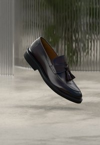 Mocassin en cuir marron foncé avec détail à pompon, texture lisse, bout arrondi et semelle en caoutchouc noire. Suspendu devant un fond texturé.