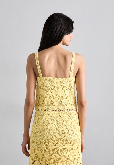 Haut en crochet jaune à bretelles spaghetti, présentant un motif floral géométrique. Le tissu a une finition texturée et est superposé.