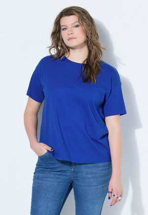 Young woman with wavy hair wears blue T-shirt and jeans, standing with one hand in pocket against white background.
