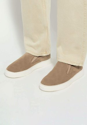 Beige suede slip-on shoes with elastic side panels, white rubber soles, and a smooth finish. Worn with light-colored jeans.