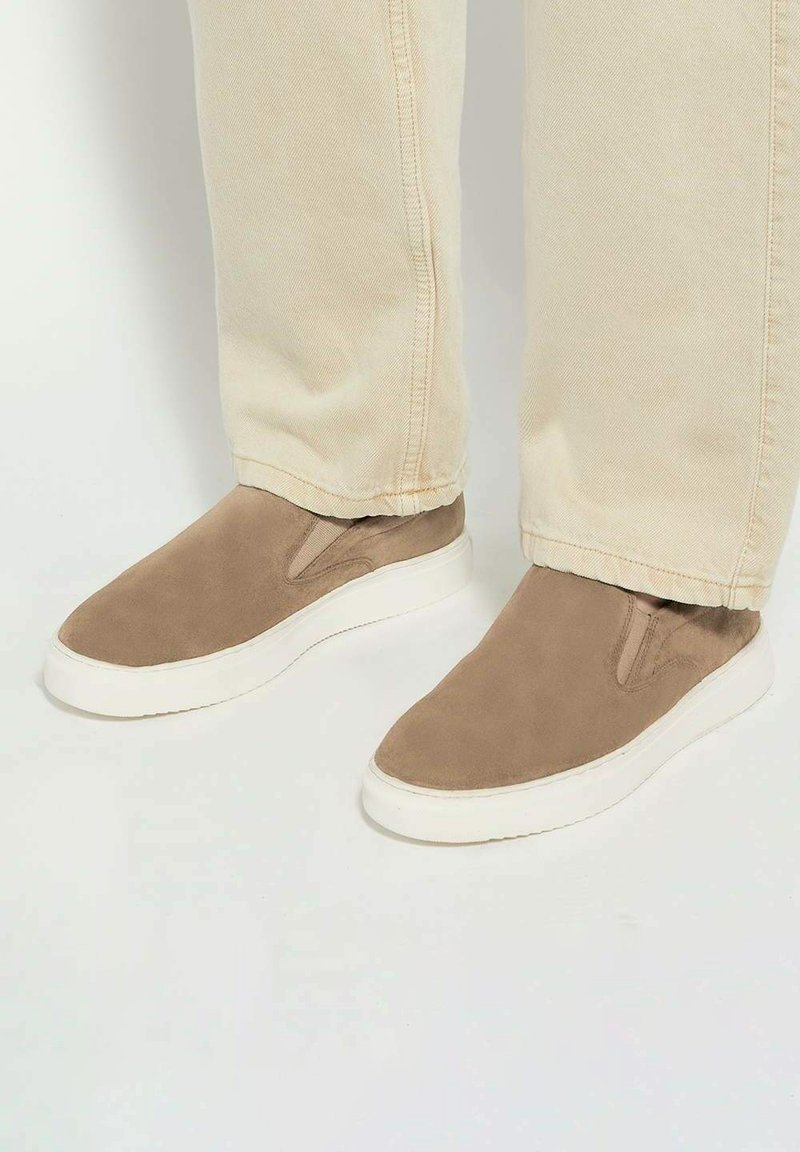 Beige suede slip-on shoes with elastic side panels, white rubber soles, and a smooth finish. Worn with light-colored jeans.