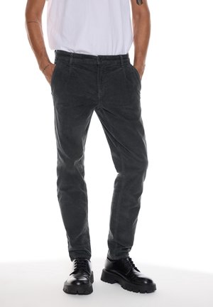 Homme portant un pantalon noir ajusté, des chaussures noires épaisses à lacets et une chemise blanche à manches courtes, debout les mains dans les poches.
