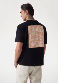 Camisa negra de manga corta con un gran parche beige que presenta patrones florales y de hojas en tonos claros, combinada con pantalones de color claro.