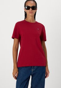 Κόκκινη βαμβακερή μπλούζα t-shirt με κλασική στρογγυλή λαιμόκοψη. Διαθέτει κοντά μανίκια και μια μικρή λεπτομέρεια λογότυπου στο κέφι. Συνδυάζεται με μπλε τζιν παντελόνι.