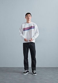 Bărbat în picioare, sprijinit de un perete gri, purtând un hanorac alb DSQUARED2, pantaloni negri și adidași alb-negru, cu mâinile ținute înainte, la talie.