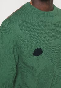 Gros plan sur un pull vert en tricot avec des motifs texturés et un seul écusson noir sur la poitrine, porté par une personne.