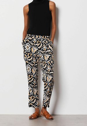 Kobieta ubrana w czarny top bez rękawów, spodnie w czarno-biały wzór z pomarańczowymi akcentami oraz brązowe płaskie sandały, stojąca z rękami w kieszeniach.