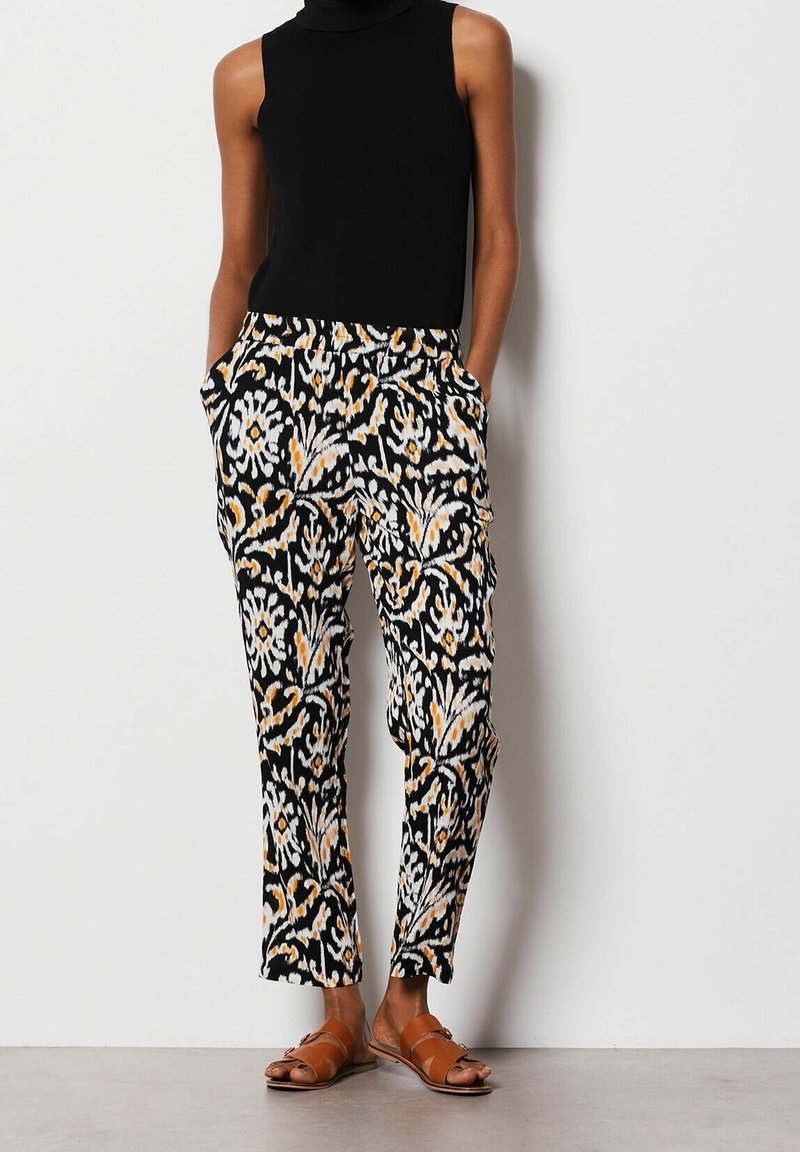 Femme portant un haut noir sans manches, un pantalon à motifs noir et blanc avec des accents orange, et des sandales plates marron, debout les mains dans les poches.
