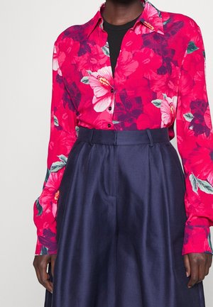 Personne portant une chemise boutonnée à fleurs rose vif rentrée dans un pantalon taille haute bleu marine sur un fond uni.