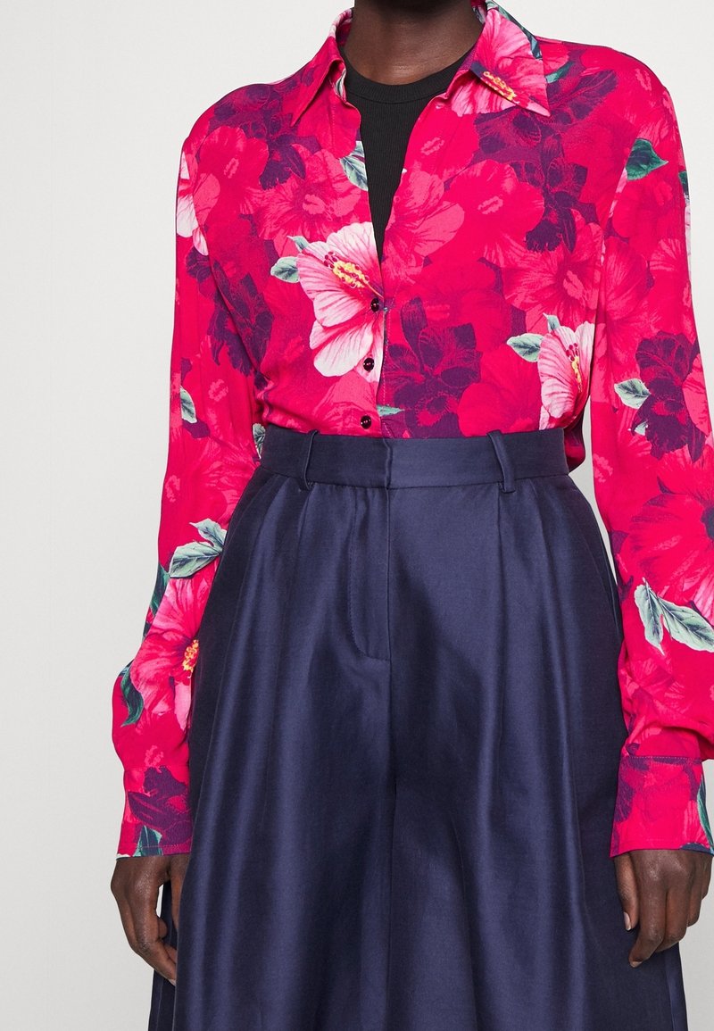 Personne portant une chemise boutonnée à fleurs rose vif rentrée dans un pantalon taille haute bleu marine sur un fond uni.