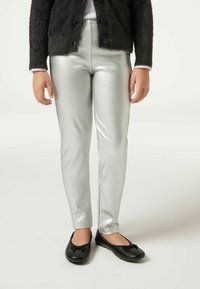 Silberne metallische Leggings mit glatter Oberfläche, haben eine schmale Passform und einen elastischen Bund. Kombiniert mit schwarzen flachen Schuhen.