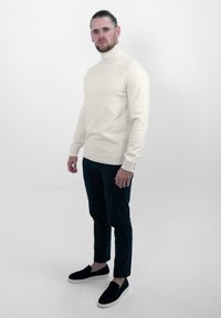 Witte coltrui met geribbelde manchetten, gecombineerd met donkerblauwe slim-fit broek en zwarte instappers. Effen achtergrond.