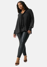 Blusa nera trasparente a maniche lunghe con collo annodato, abbinata a leggings aderenti in ecopelle nera e tacchi aperti neri.