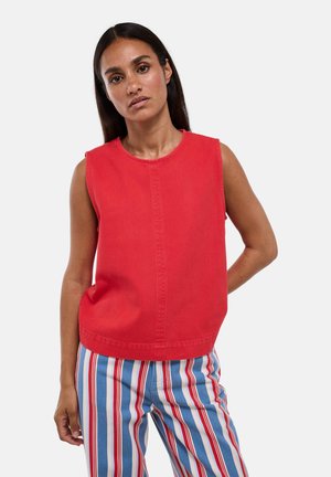 Femme aux longs cheveux foncés portant un haut rouge sans manches et un pantalon rayé verticalement bleu, rouge et blanc sur fond uni.