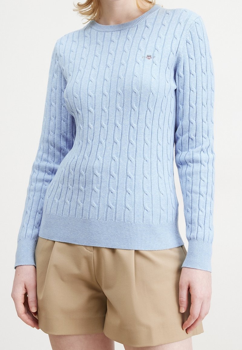 Jasnoniebieski sweter dziany z warkoczowym wzorem, okrągłym dekoltem oraz ściągaczami na mankietach i u dołu. Małe logo na piersi. W zestawie z beżowymi szortami.