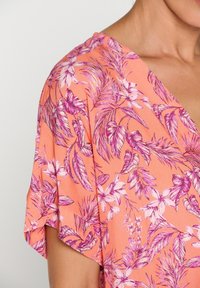 Blusa floreale color corallo con motivo di foglie e fiori rosa. Presenta una scollatura a V e maniche corte. Tessuto leggero con una consistenza liscia.