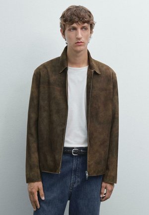 Veste en suède marron avec une coupe droite, fermeture éclair à l'avant et deux poches poitrine, portée sur un t-shirt blanc et un jean en denim bleu.