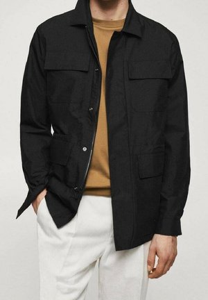 Mężczyzna w czarnej kurtce na zamek i guziki, pod którą ma brązową koszulę, białe spodnie, ręka w kieszeni, stojący na jednolitym tle.