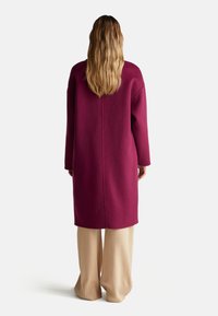 Eine Person mit langen, welligen Haaren, die einen langen magentafarbenen Mantel und lockere beige Hosen trägt, steht mit dem Rücken zu einer weißen Hintergrund.