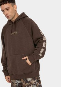 Brązowa bluza z kapturem z raglanowymi rękawami, przednią kieszenią i delikatnymi akcentami z logo na rękawach. Miękka w dotyku i luźnego kroju.