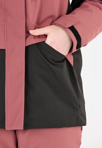Vattentålig jacka med en rosa överdel och svart underdel, med en texturerad tyg och en sidoficka. Kardborreakcenter vid manschetten.