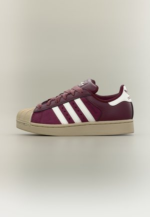 Baskets Adidas bordeaux avec trois bandes blanches, semelle beige et embout, lacets bordeaux et languette de talon blanche avec logo.