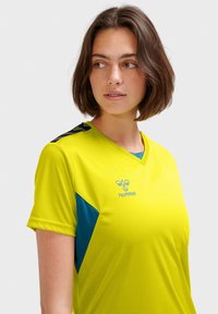 Ljusgul atletisk tröja med korta ärmar, med en blå accent på sidan och en svart randig detalj på axeln. Hummel-logotypen finns.