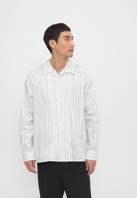 Camisa blanca de manga larga con delgadas rayas verticales azules, cuello puntiagudo y cierre de botones, con un corte holgado y mangas con puños.