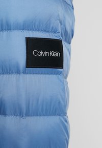 Jasnoniebieska pikowana kamizelka o błyszczącej fakturze, z czarną prostokątną metką z napisem "Calvin Klein" w białych literach.