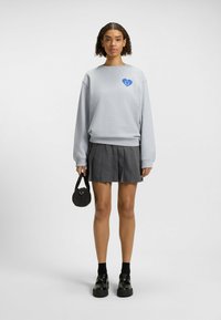 Grijze sweatshirt met een blauwe hart-smiley graphic, gecombineerd met een geplooide grijze rok, zwarte ronde handtas en chunky zwarte schoenen.