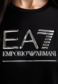 Melna kokvilnas T-kreklā ar metāliskām sudraba krāsas logo "EA7" un "EMPORIO ARMANI", ar gludu, spīdīgu tekstūru un raksturīgām dizaina iezīmēm.