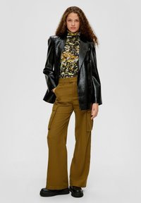 Zwarte faux leren blazer, geprinte coltrui met gele en blauwe accenten, en olijfgroene wijde cargo broek. Chunky zwarte laarzen.